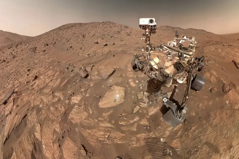 Rover Perseverance em Marte analisando rochas em uma região montanhosa e seca da superfície marciana.